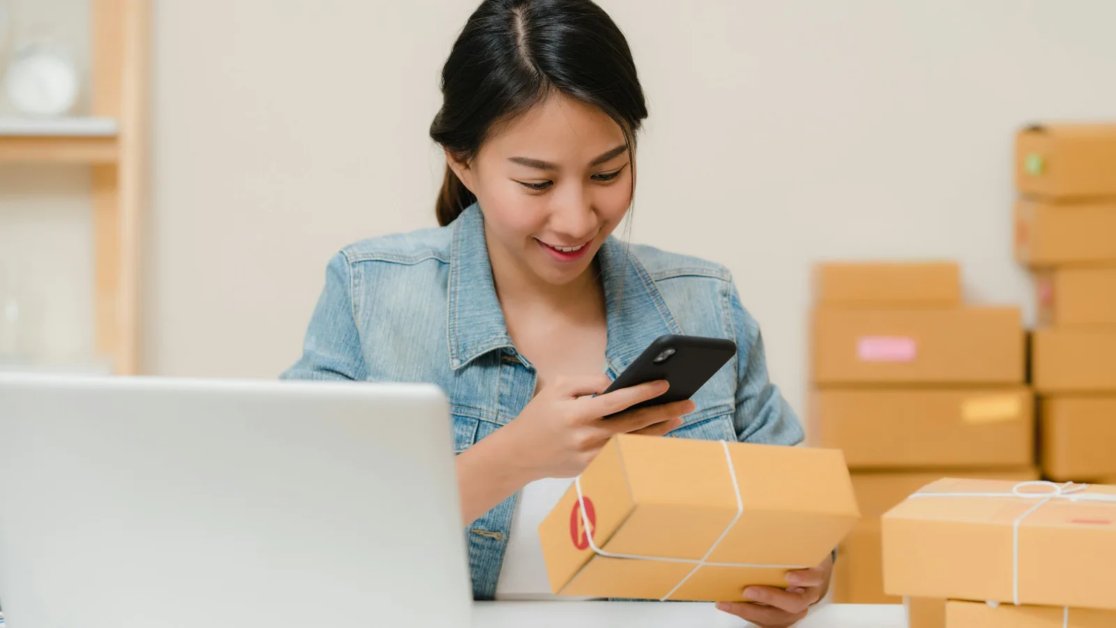 A woman at a laptop takes a photo of a packed box with her phone, with more boxes stacked behind her