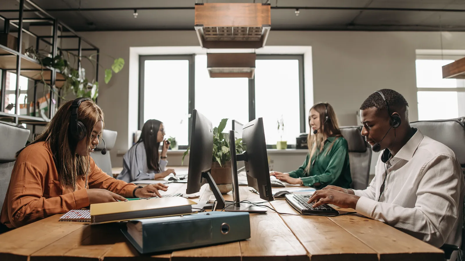 Four people wearing headsets type on laptops side by side at a long wooden table in a bright office
