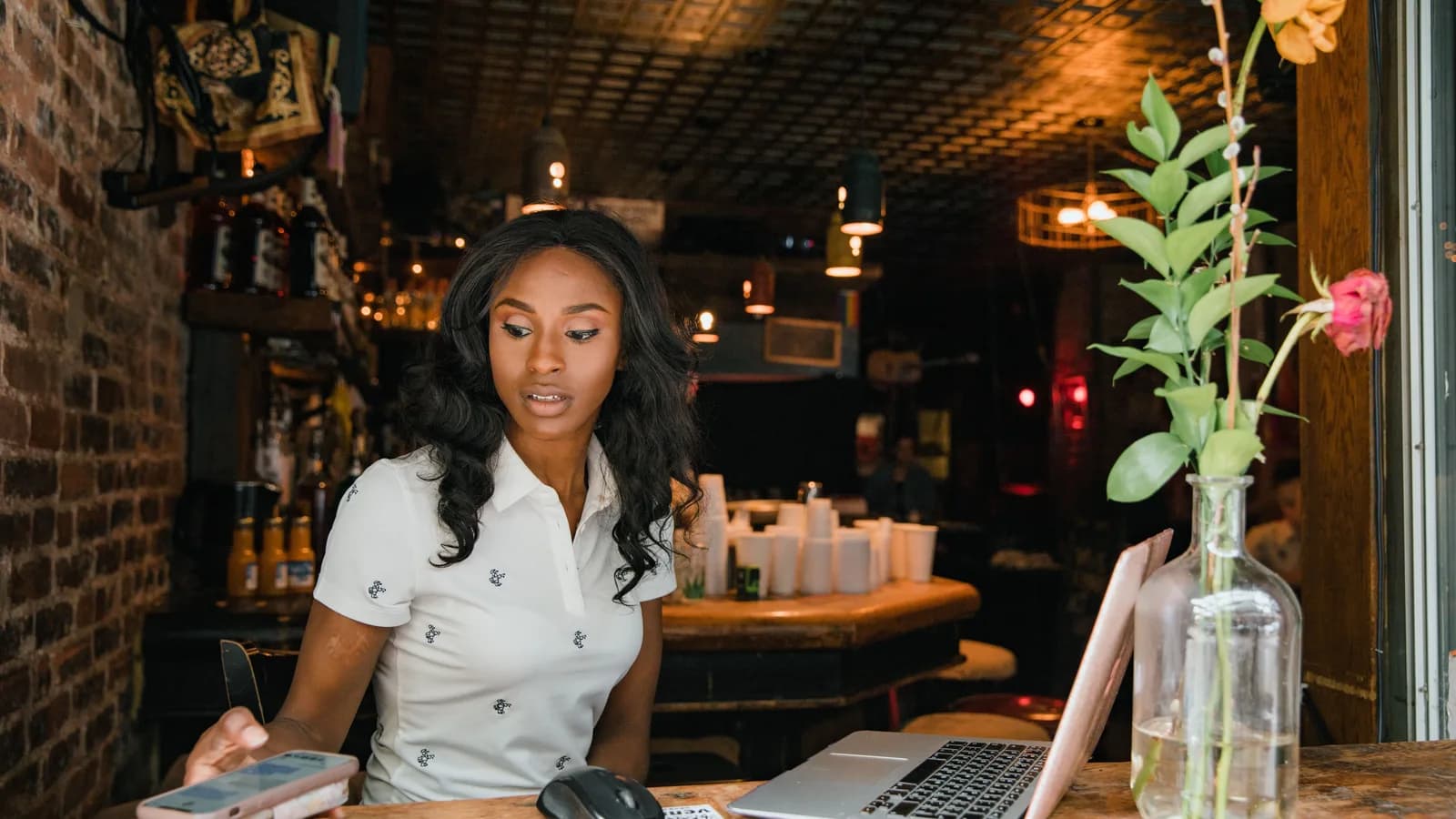 A woman at a cafe table reads a message on her phone with an open laptop beside her