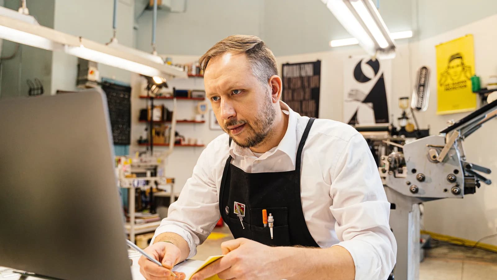 Man in a work apron checking a laptop and jotting notes next to a large printing press
